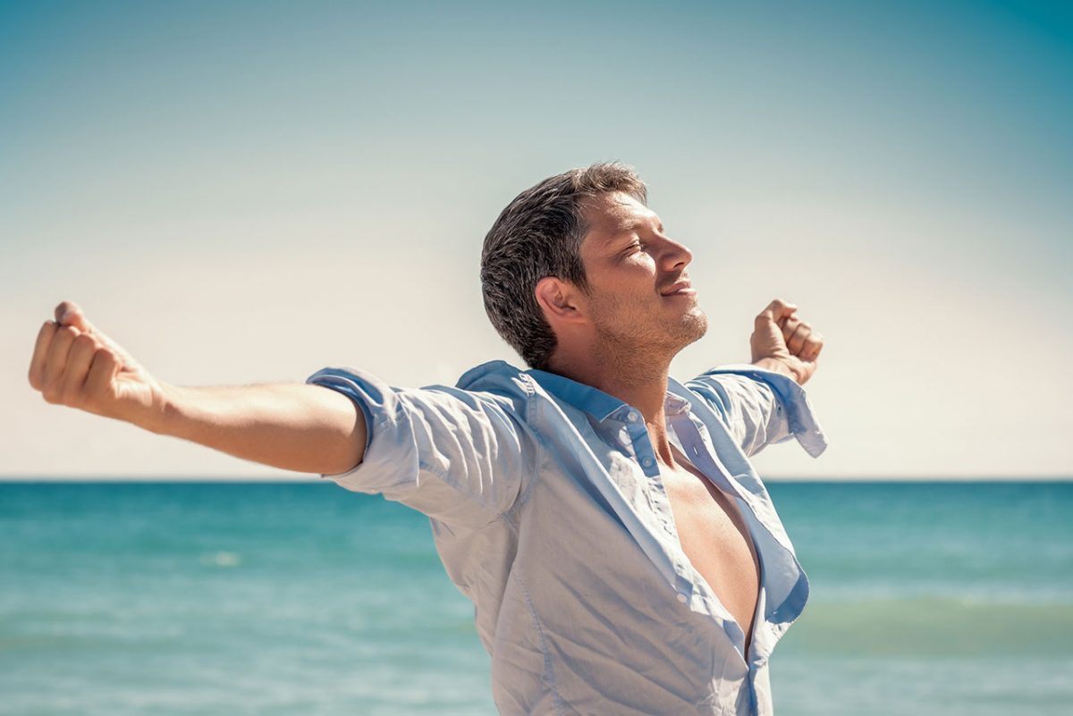 Mann steht am Meeresstrand, breitet die Arme weit aus und atmet tief durch